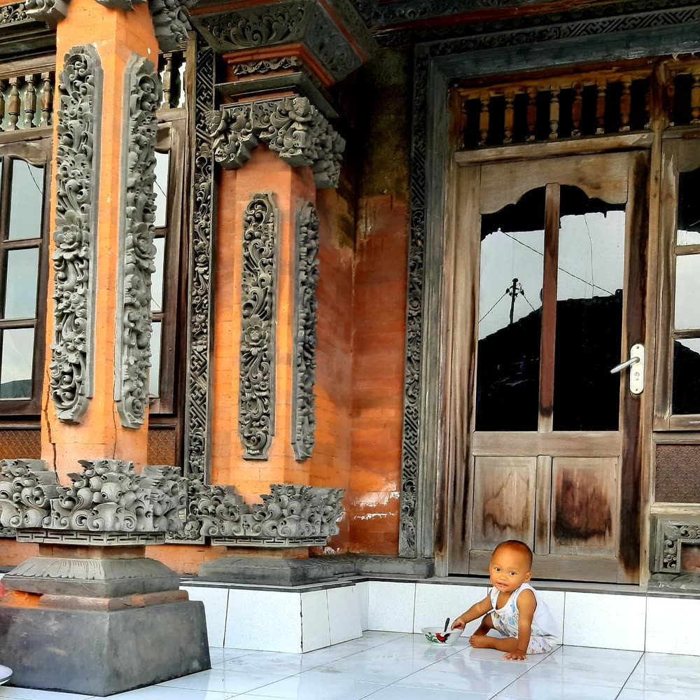 Carved stone and brick facade of a traditional Balinese family compound, a small child playing on the terrace